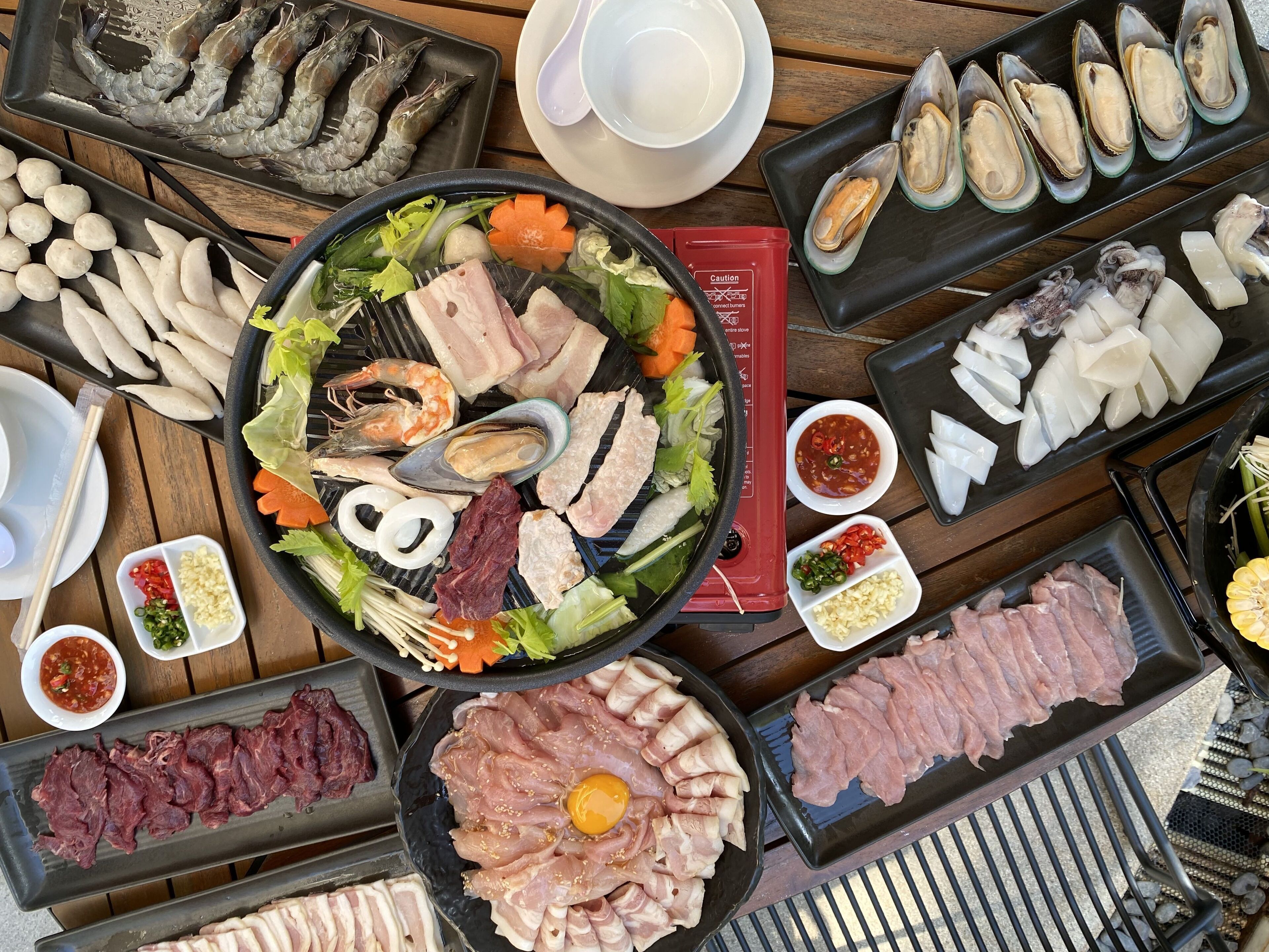 A variety of raw seafood and meats arranged on a wooden table, including shrimp, oysters, and different cuts of meat.