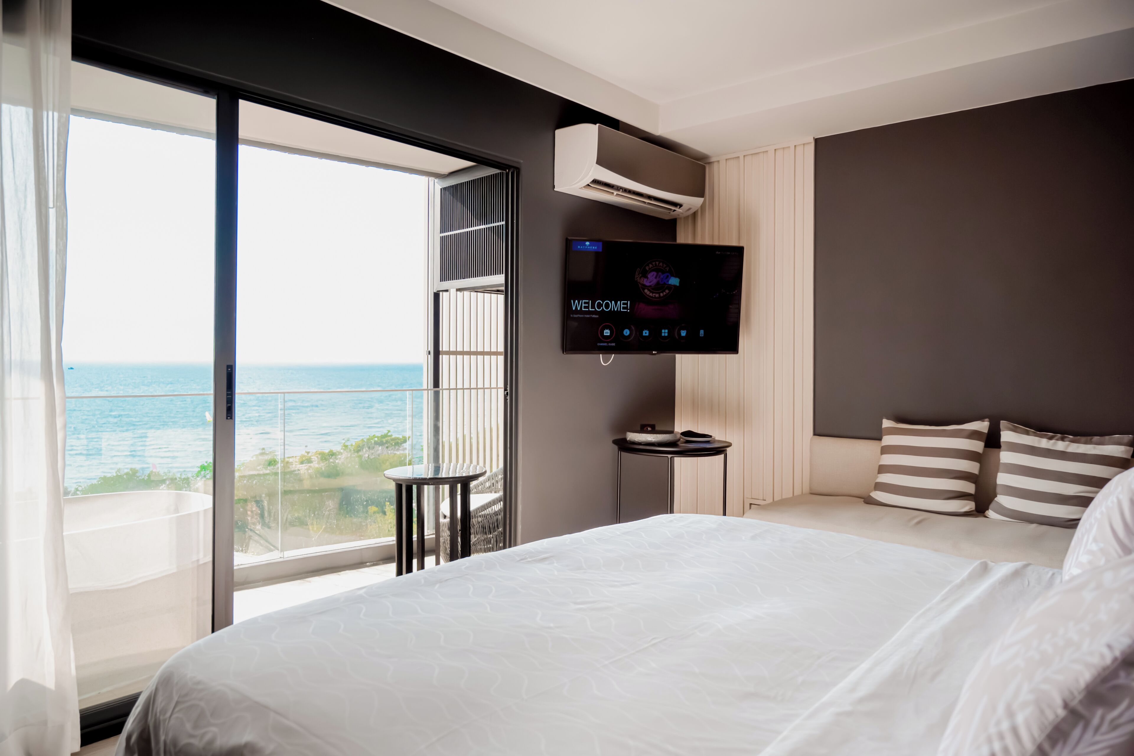 A hotel room with a large bed, a TV mounted on the wall, a small table, and a balcony with a view of the sea.
