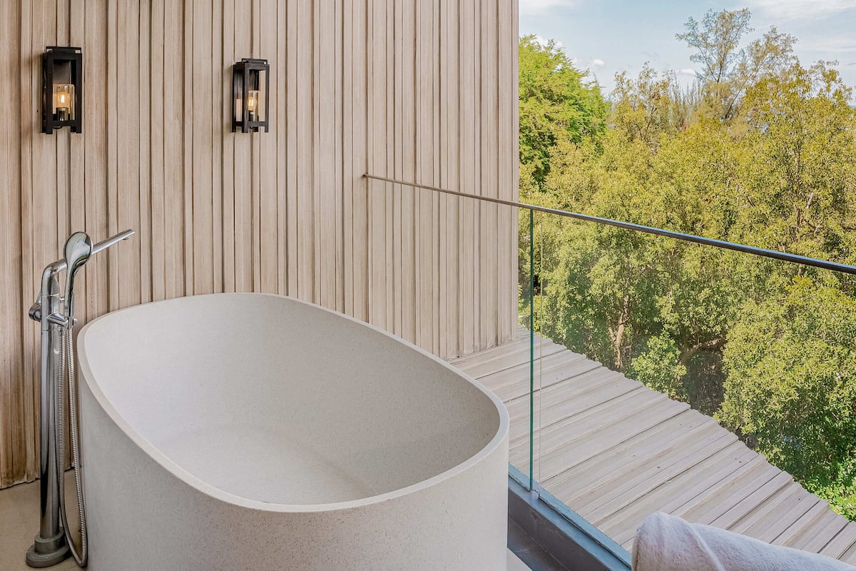 Ein modernes Badezimmer mit einer freistehenden Badewanne, Holzpaneele und einem Glasgeländer mit Blick auf ein bewaldetes Gebiet.