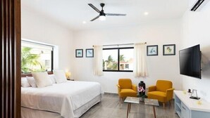 A hotel room with a bed, two yellow armchairs, a small table, a TV, and a ceiling fan.