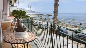 A balcony with a potted tree, a tiled table, and a view of the sea and rocky coastline. - SUN SEA HOUSE TAORMINA  (Giardini Naxos)