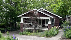 Ein zweistöckiges Haus mit Veranda, umgeben von üppigem Grün und blühenden Pflanzen.