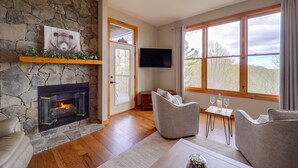 A cozy living room with a stone fireplace, comfortable seating, a flat-screen TV, and large windows offering a view of the outdoors. - Hike & Ski: Mtn-View Gem w/ Balcony in Burnsville! (Burnsville)
