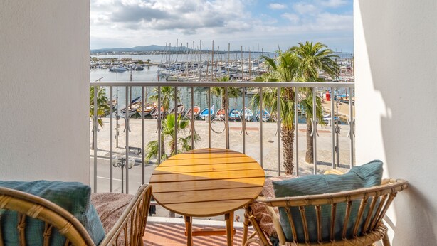 A balcony with a wooden table and wicker chairs, overlooking a marina with boats and palm trees.
