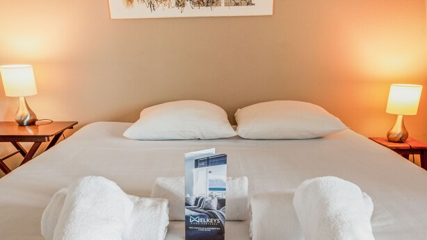 Una cama bien hecha con sábanas blancas, dos almohadas y un juego de toallas dobladas al pie. Hay dos mesitas de noche con lámparas, una obra de arte enmarcada en la pared y un folleto sobre la cama.