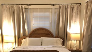 A bedroom with a large bed, a wicker sofa with cushions, a nightstand with a lamp, and a window with blinds. - Convenient quiet LSU Location near City Park, coffee shops, restaurants and bars (Baton Rouge)