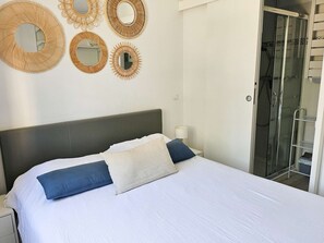 A bedroom with a bed, pillows, a bedside table, a mirror, and a shower area.