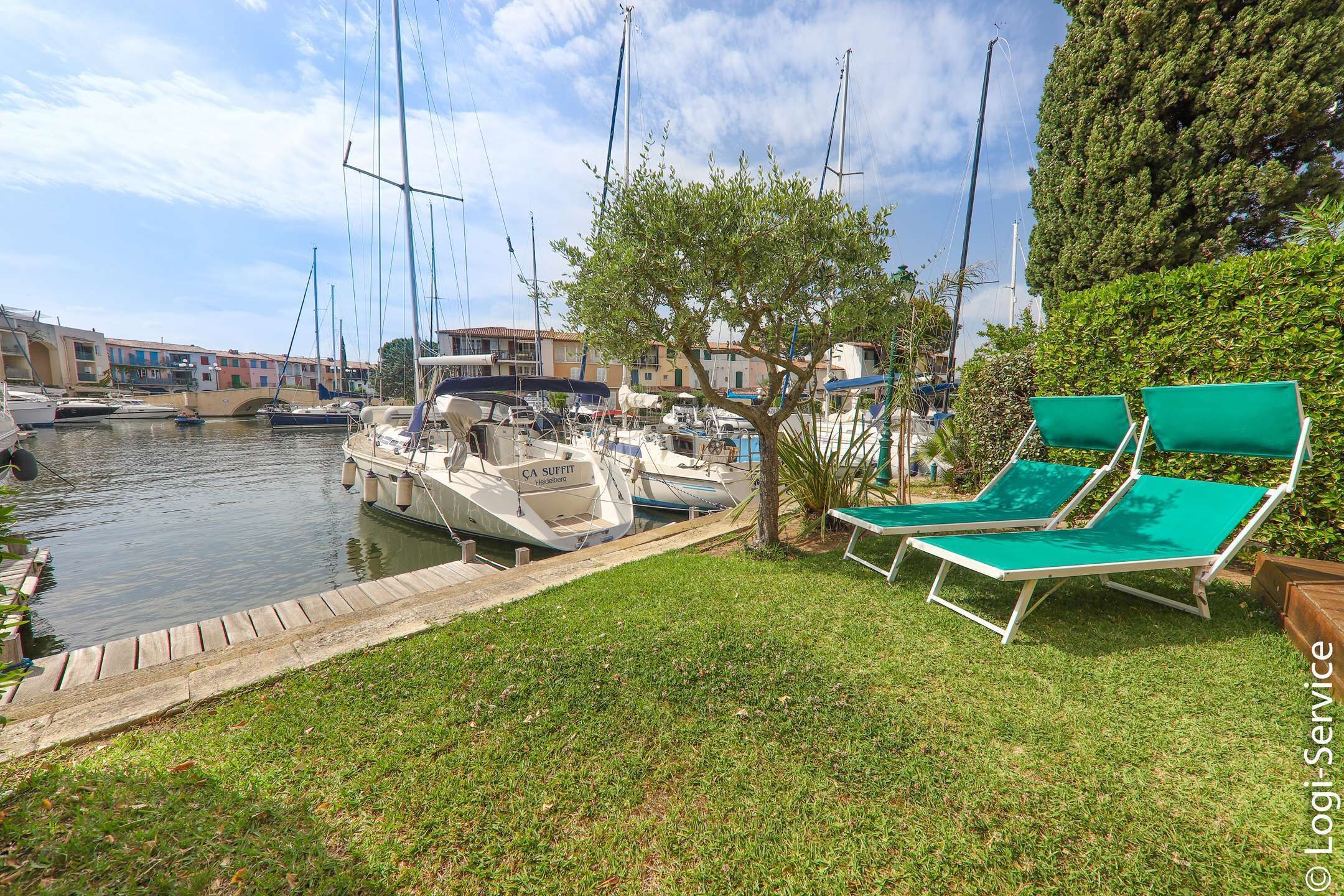 Un puerto deportivo con barcos amarrados, un área verde con sillones de descanso y edificios al fondo.