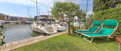 Un puerto deportivo con barcos amarrados, un área verde con sillones de descanso y edificios al fondo.