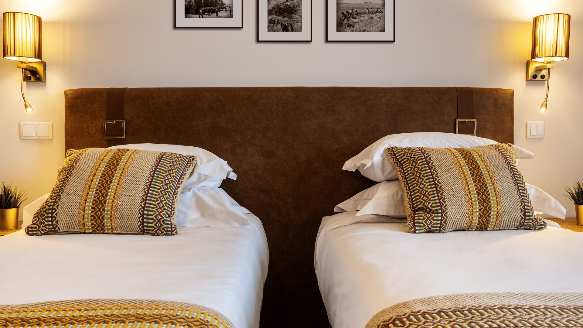 A hotel room with two beds, each with patterned pillows and a brown headboard. There are wall-mounted lamps on either side of the bed.