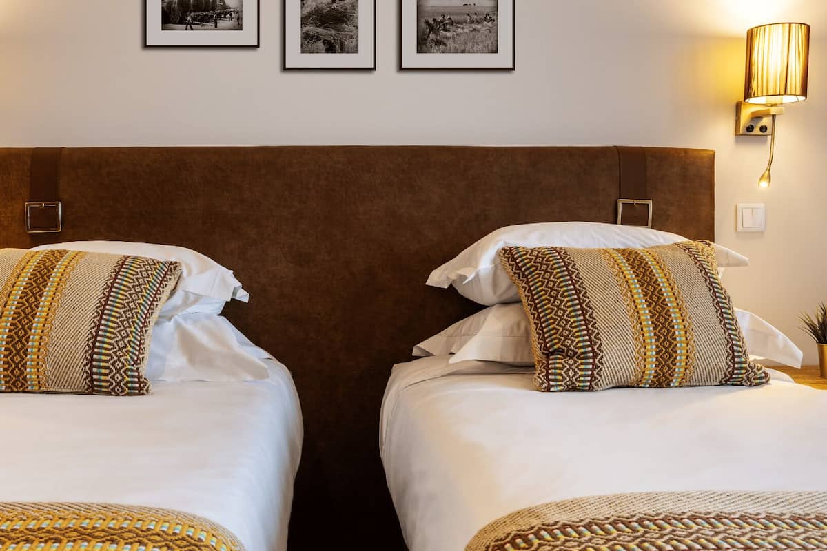 Ein Hotelzimmer mit zwei Betten, jeweils mit gemusterten Kissen und einem braunen Kopfteil. An jeder Seite des Bettes befinden sich Wandlampen.