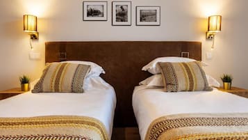 A hotel room with two beds, each with patterned bedding and pillows, and wall-mounted lamps.