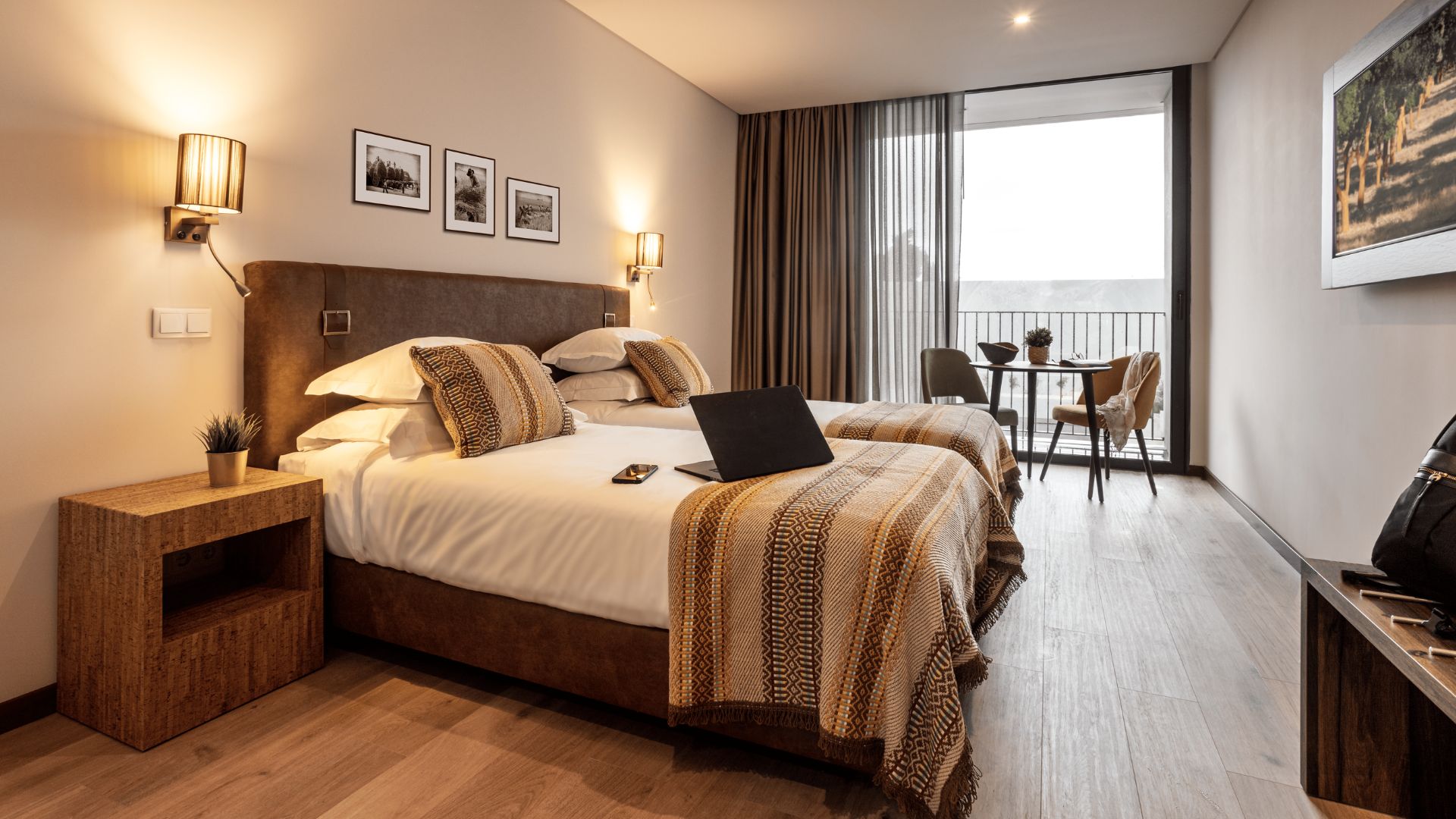 A hotel room with a large bed, a desk with a laptop, a small table with chairs, and a balcony with curtains.