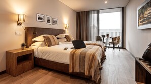 A hotel room with a large bed, a desk with a laptop, a small table with chairs, and a balcony with curtains.