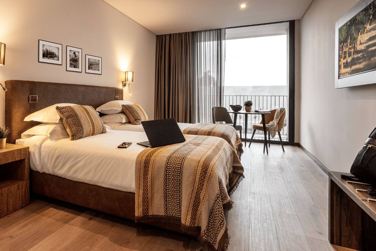 Ein Hotelzimmer mit einem großen Bett, einem Schreibtisch mit einem Laptop, einem kleinen Tisch mit Stühlen und einem Balkon mit Vorhängen.