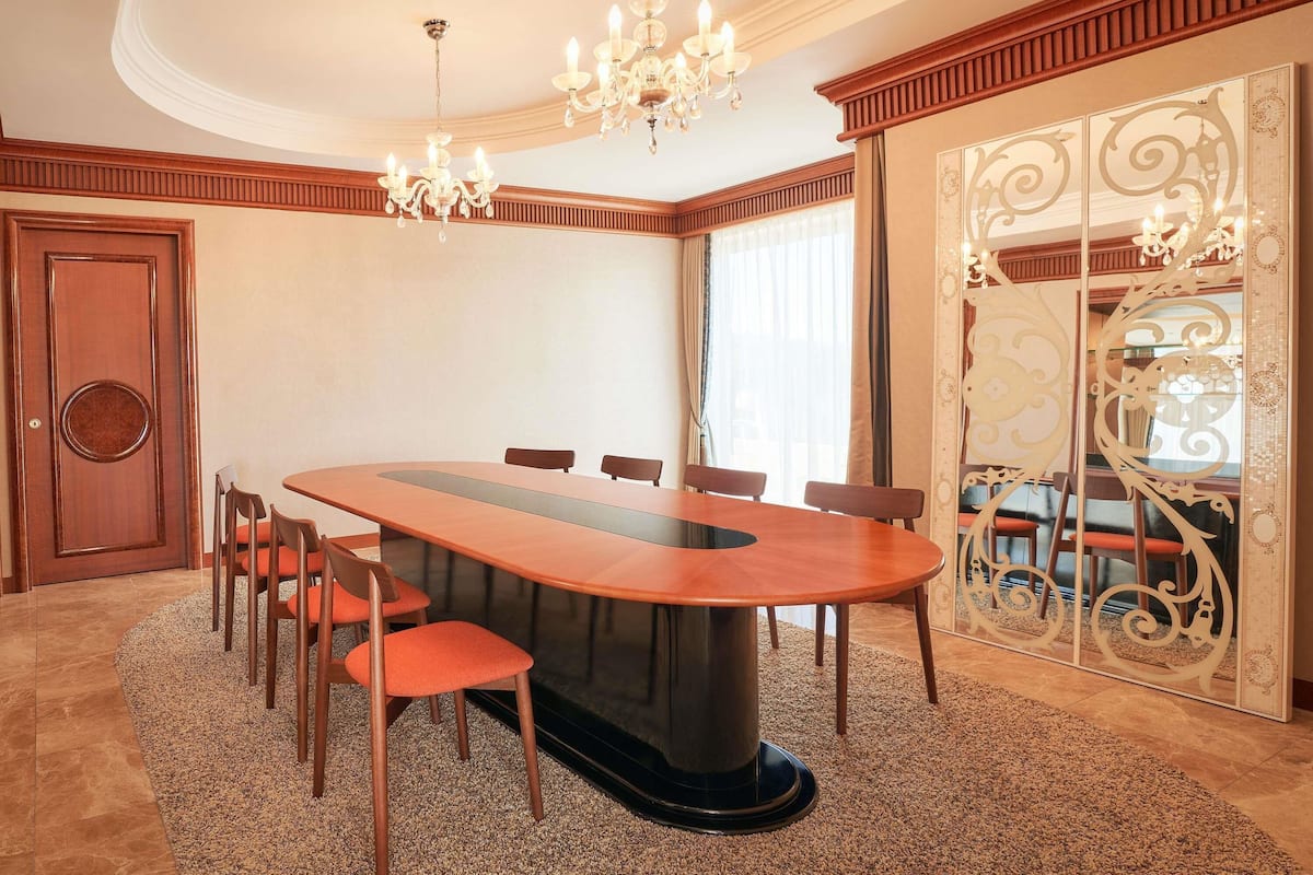Un salón de conferencias con una larga mesa, sillas, una araña y un espejo decorativo.