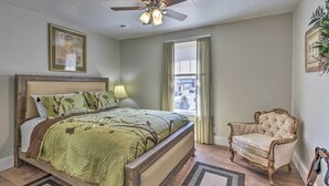 Un dormitorio con cama, una silla, ventilador de techo, ventana con cortinas y un cuadro en la pared.