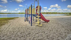 Un parque infantil con toboganes y estructuras para escalar en una playa arenosa, con un lago al fondo.