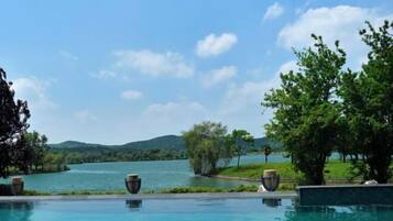 Una piscina con vistas a un lago y colinas, rodeada de árboles y mobiliario exterior.