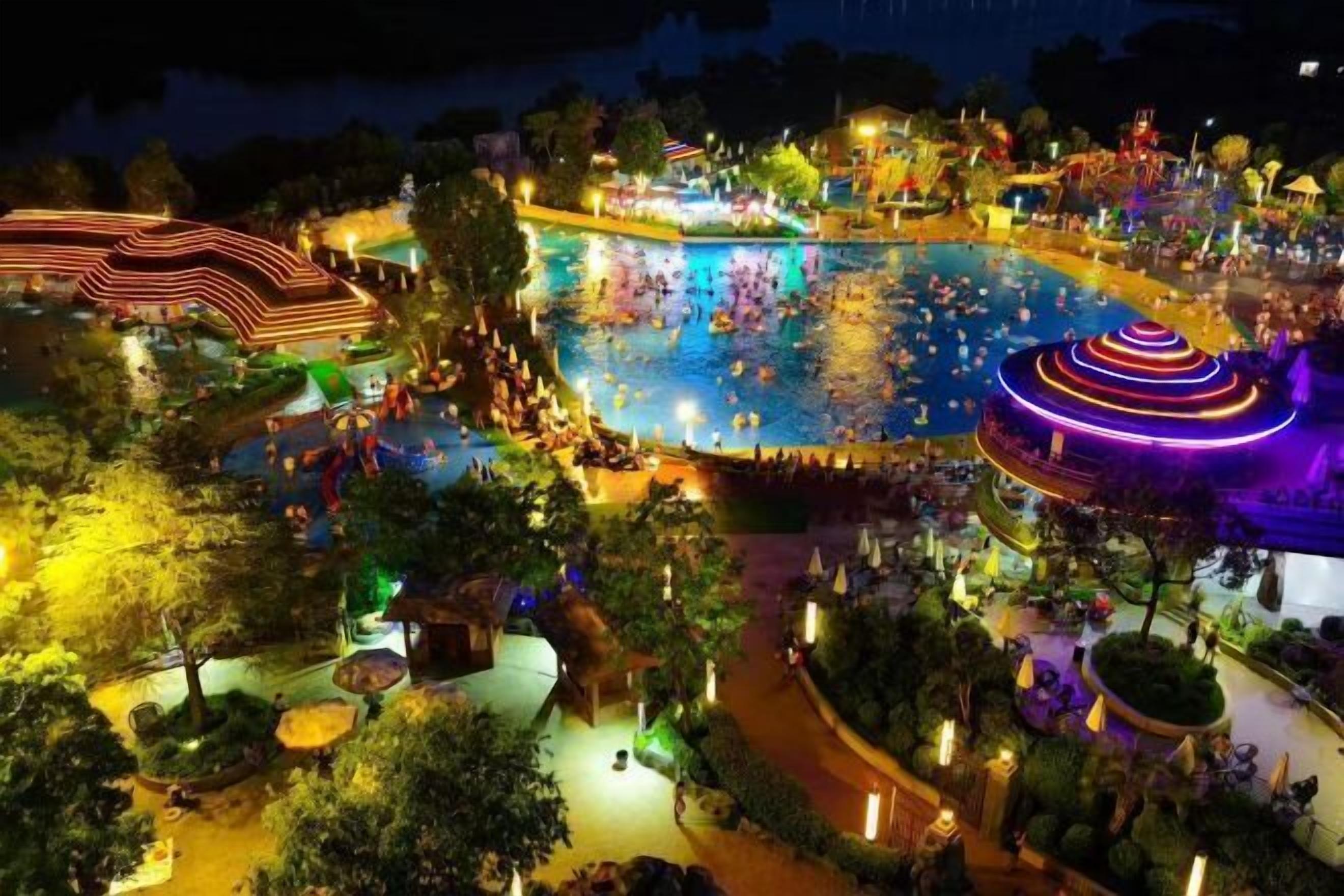A large outdoor swimming pool surrounded by trees and pathways, with people swimming and walking around.
