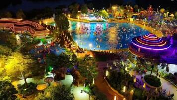 A large outdoor swimming pool surrounded by trees and pathways, with people swimming and walking around.
