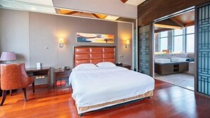 Habitación de hotel moderna con una cama grande, suelo de madera, un escritorio con lámpara y un ventanal con vistas al exterior.