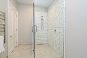 Pequeño espacio de ducha cerrado, con paredes y suelo de azulejos blancos, puerta de cristal y un perchero con una toalla blanca.