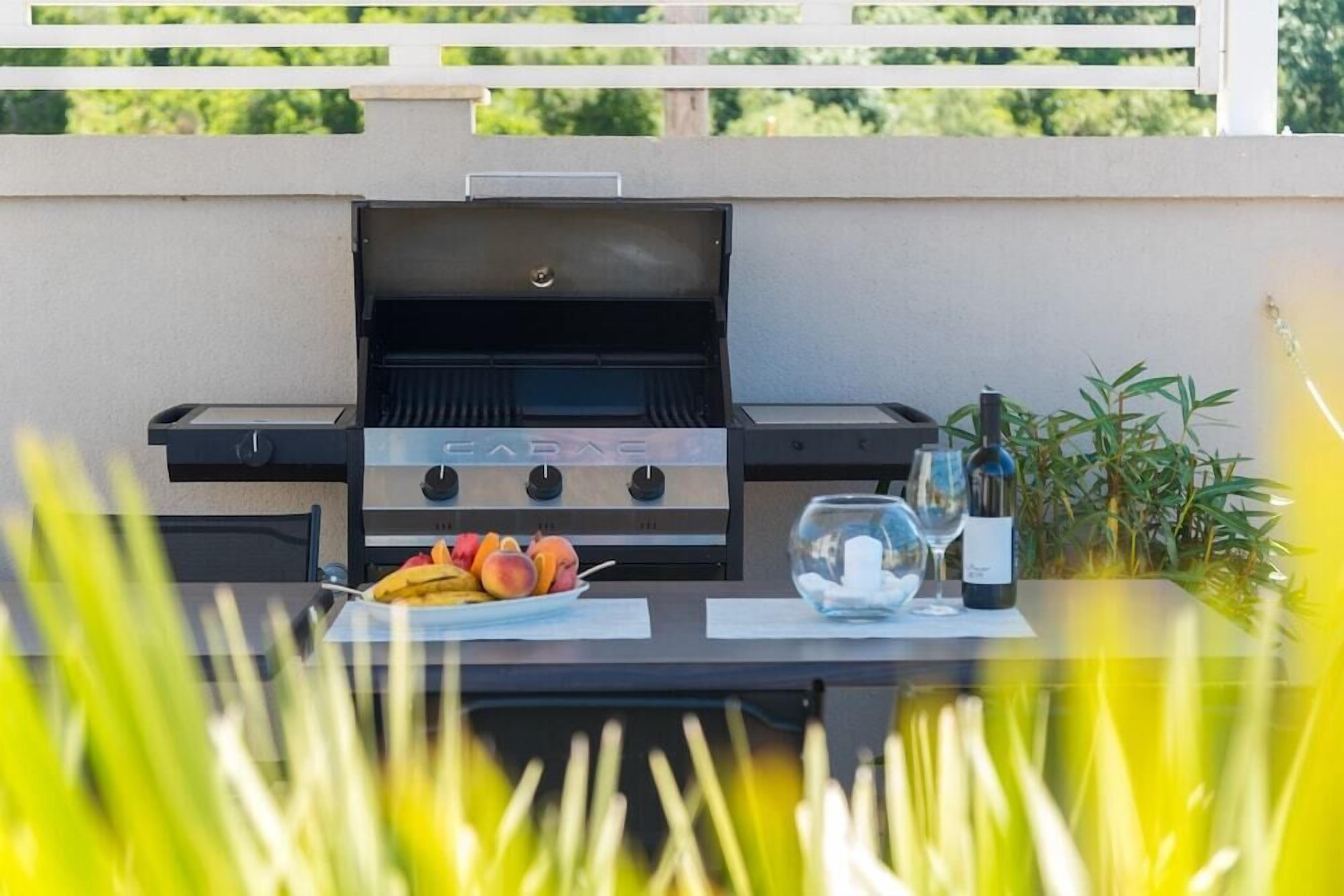 An outdoor grill with a table set for a meal, including fruits and a bottle of wine.