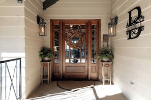 Une porte d’entrée en bois ornée d’une guirlande, de deux plantes en pot et d’une enseigne fixée au mur.