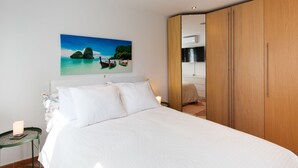 Een slaapkamer met een bed, een schilderij van een tropisch landschap, een houten garderobe en een klein tafeltje met een lamp.