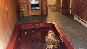 Interior de una cabaña de madera con un jacuzzi en el centro, una puerta que da a la cocina y piso de baldosas.