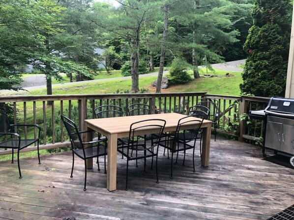 A wooden deck with a table and chairs, a grill, and a view of a wooded area.