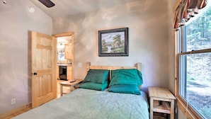 A bedroom with a bed, green pillows, a wooden headboard, a nightstand, a small table, a window with a view of trees, and a painting on the wall.