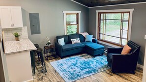 Ein modernes Wohnzimmer mit Küche, einem blauen Ecksofa, einem kleinen Beistelltisch und einem großen Fenster.