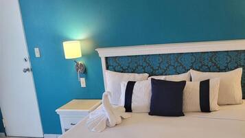 Habitación de hotel con una pared verde azulado, una cama blanca con varias almohadas, una lámpara de noche y una toalla blanca con forma de cisne.