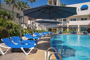 A poolside area with blue lounge chairs and umbrellas, surrounded by palm trees and a building with large windows. - Sunset Palace Beach Hotel (Mazatlán)