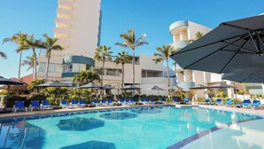 A swimming pool with blue water, surrounded by lounge chairs and umbrellas, with palm trees and modern buildings in the background. - Sunset Palace Beach Hotel (Mazatlán)