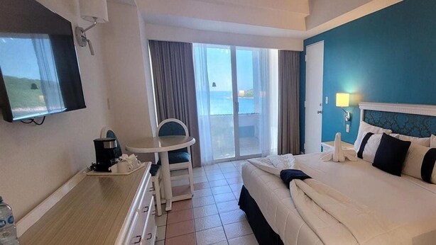A hotel room with a bed, a desk, a chair, a TV, and a view of the sea through the window. - Sunset Palace Beach Hotel (Mazatlán)