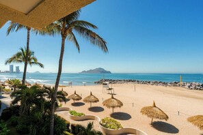 A beach with palm trees, thatched umbrellas, and a view of the ocean. - Sunset Palace Beach Hotel (Mazatlán)