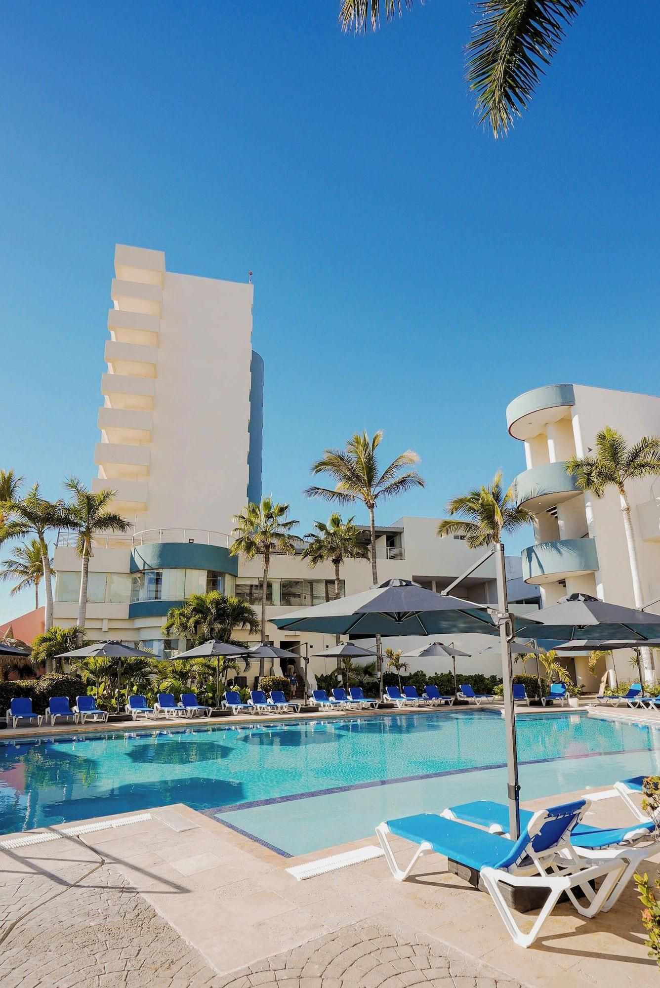 Un área de piscina con sillones reclinables y sombrillas, rodeada de palmeras y edificios modernos.