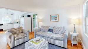 A modern living room with striped sofas, a coffee table, and a kitchen in the background. - Charming Chatham Cottage  (Chatham)