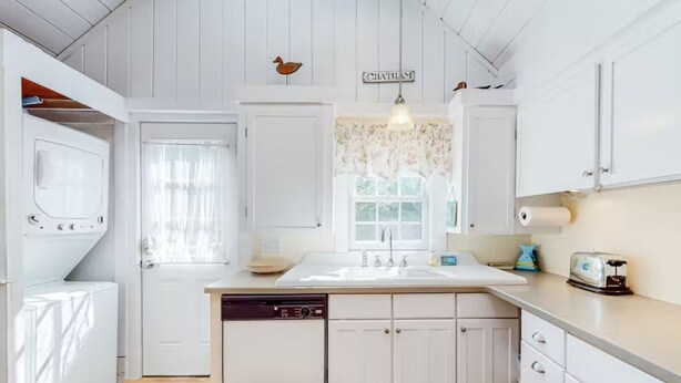 A kitchen with white cabinets, a dishwasher, a sink, and a window with a floral curtain. - Charming Chatham Cottage  (Chatham)