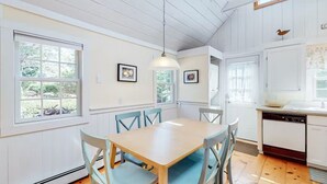 A dining area with a wooden table and blue chairs, a hanging light fixture, and a kitchen with white cabinets and a dishwasher. - Charming Chatham Cottage  (Chatham)