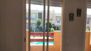A view through a sliding glass door to an outdoor area with a swimming pool, a red fence, and buildings in the background.