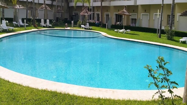 Una piscina rodeada de sillones de descanso y sombrillas, con un edificio de varios pisos al fondo.