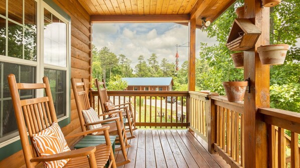 A wooden porch with several rocking chairs and a view of a cabin in the distance.