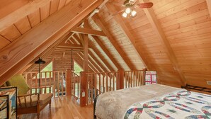 Une chambre mansardée chaleureuse avec un plafond en bois, un lit recouvert d’une courtepointe, une chaise et une bibliothèque.