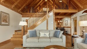 Un vaste salon doté d’un haut plafond en bois, d’un escalier, d’un canapé avec des coussins et, au fond, d’un coin repas.