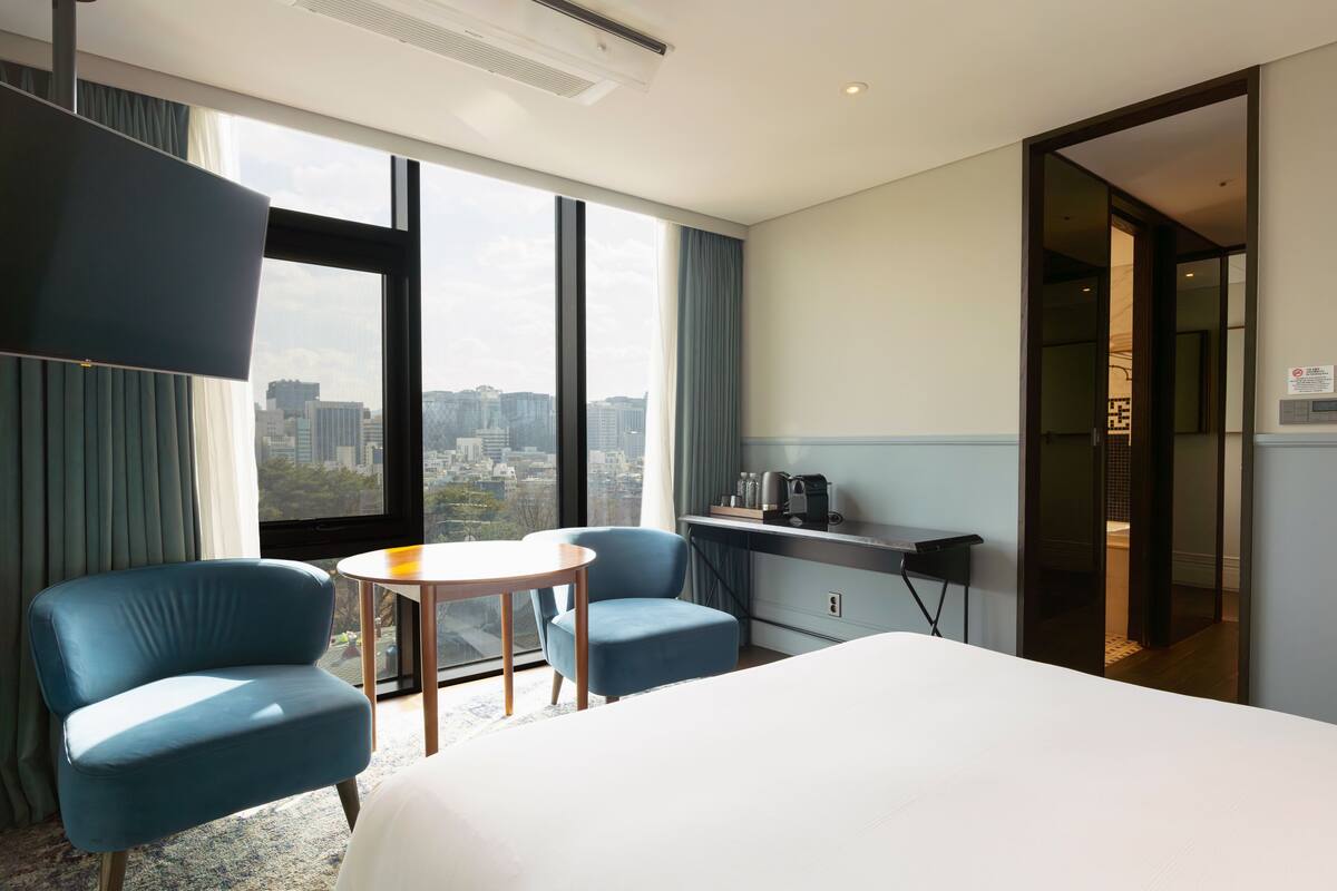 A hotel room with a bed, two armchairs, a small table, a desk with a coffee maker, and a view of the city through large windows.