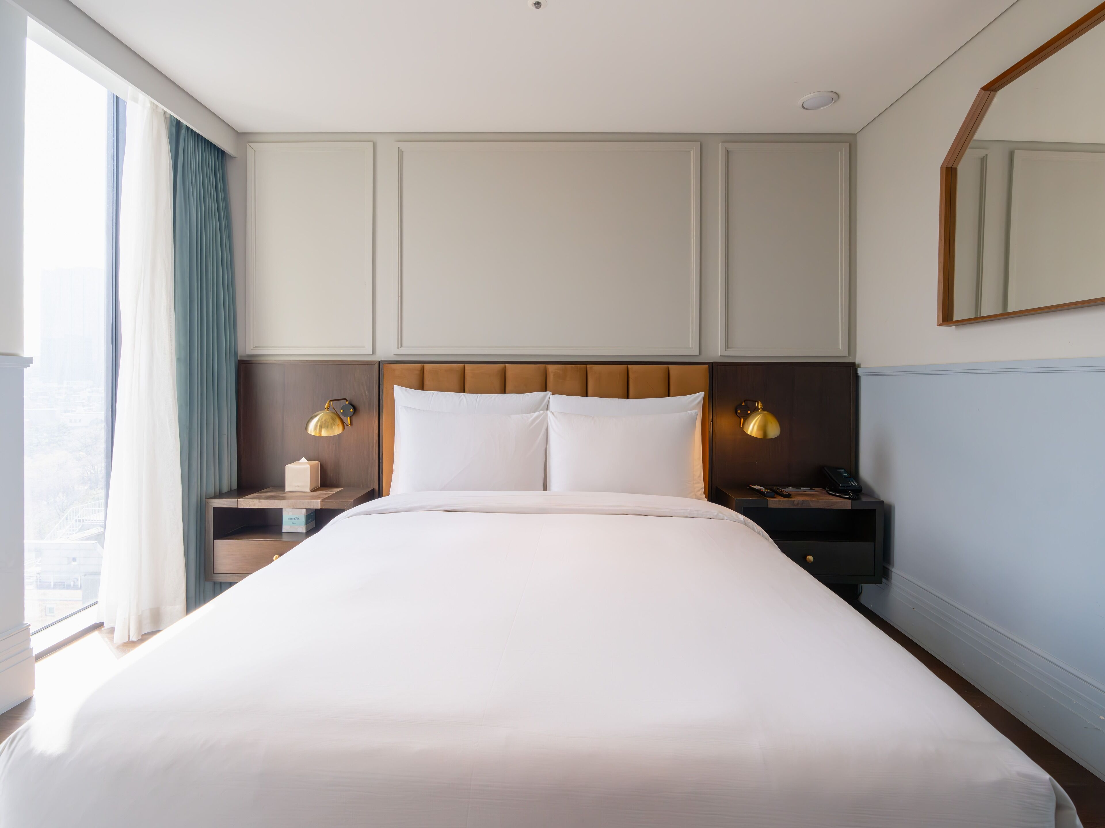 Une chambre d’hôtel avec un grand lit, deux tables de chevet, une commode, un miroir et une fenêtre avec des rideaux.